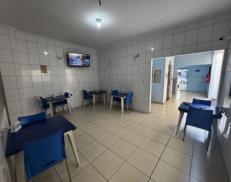 Uma sala de jantar com mobília esparsa, paredes revestidas de azulejos brancos, cadeiras azuis e mesas quadradas dispostas sobre um piso de azulejos claros.