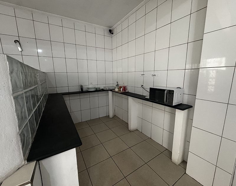 Uma cozinha minimalista com paredes e pisos revestidos com azulejos brancos, bancada preta e micro-ondas em uma prateleira.
