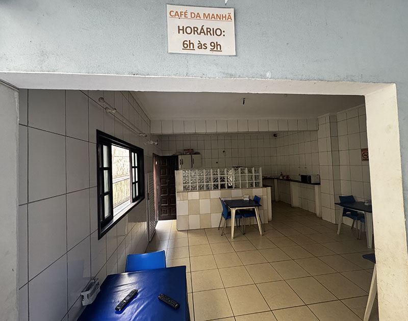 Uma área de refeições simples, com paredes revestidas de azulejos e mesas com toalhas azuis, com uma placa indicando que o café da manhã é servido das 6h às 9h.