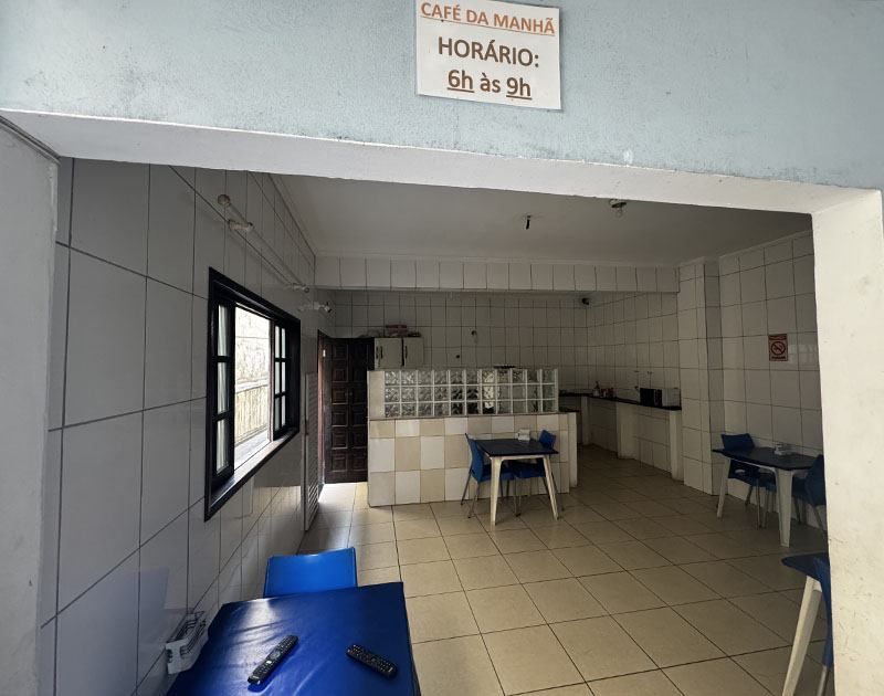 Sala de café da manhã de hotel com paredes revestidas de azulejos brancos, mesas e cadeiras azuis e uma placa indicando que o horário do café da manhã é das 6h às 9h.
