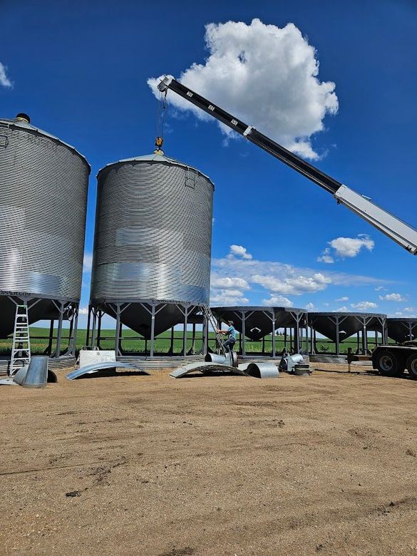 Large silver grain silos being lifted by a crane under a blue sky at an industrial site