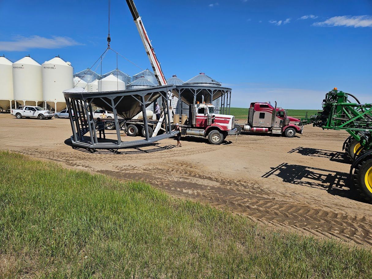 Farm equipment and a crane lifting a large metal structure in a rural field beside silos.