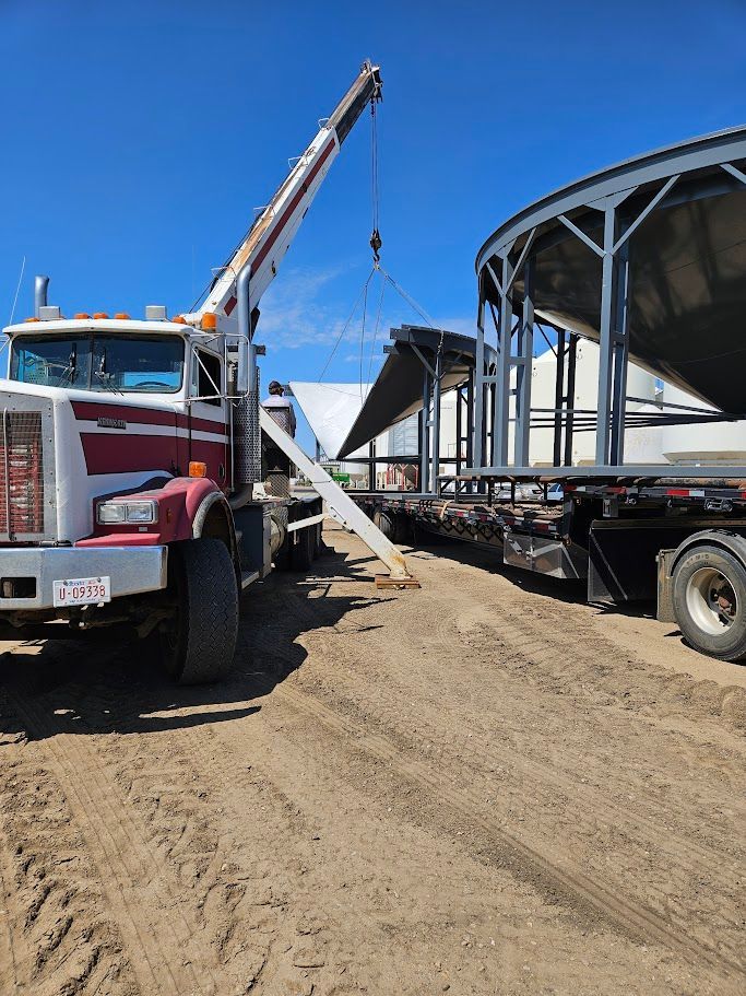 Crane loading a large curved steel structure onto a trailer in a dirt lot