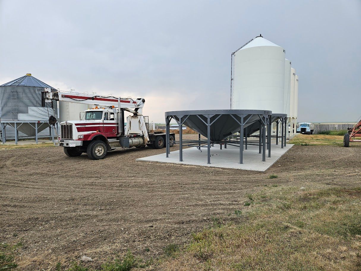 Red and white truck beside farm silos and grain bins in a rural field.