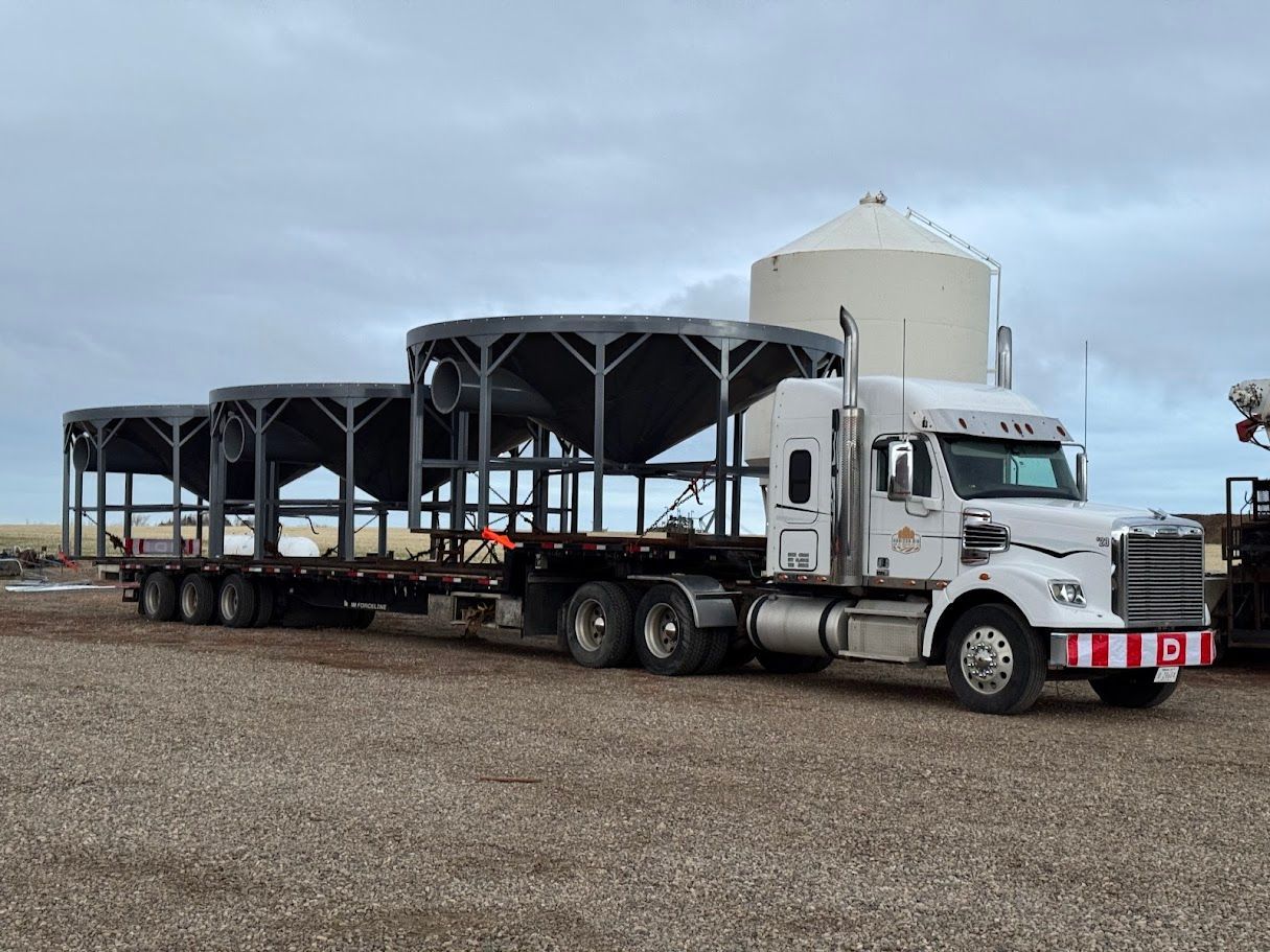White semi-truck hauling a long black metal structure in a gravel lot under cloudy sky
