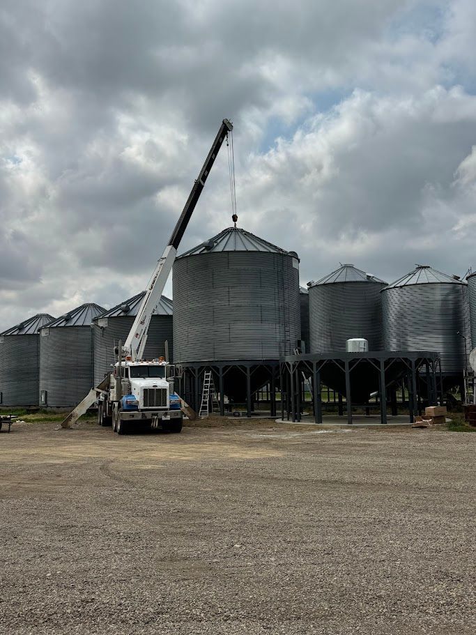 Crane lifting a grain bin among metal silos under a cloudy sky