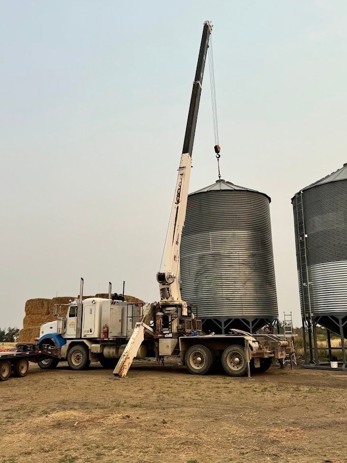 Crane truck lifting or lowering a large metal grain bin in a rural field near other silos.