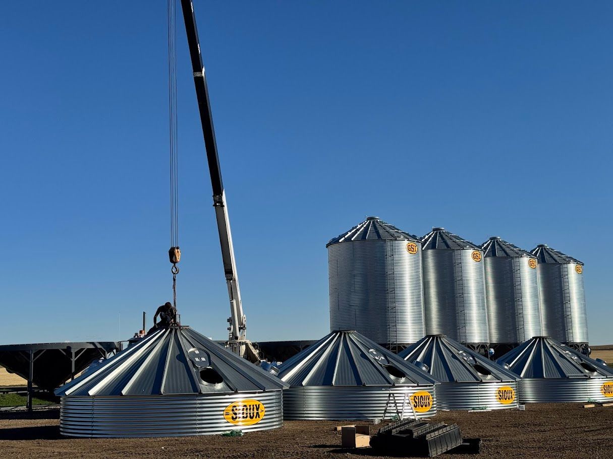 Crane lifting corrugated metal grain bins in a farmyard under a clear blue sky.