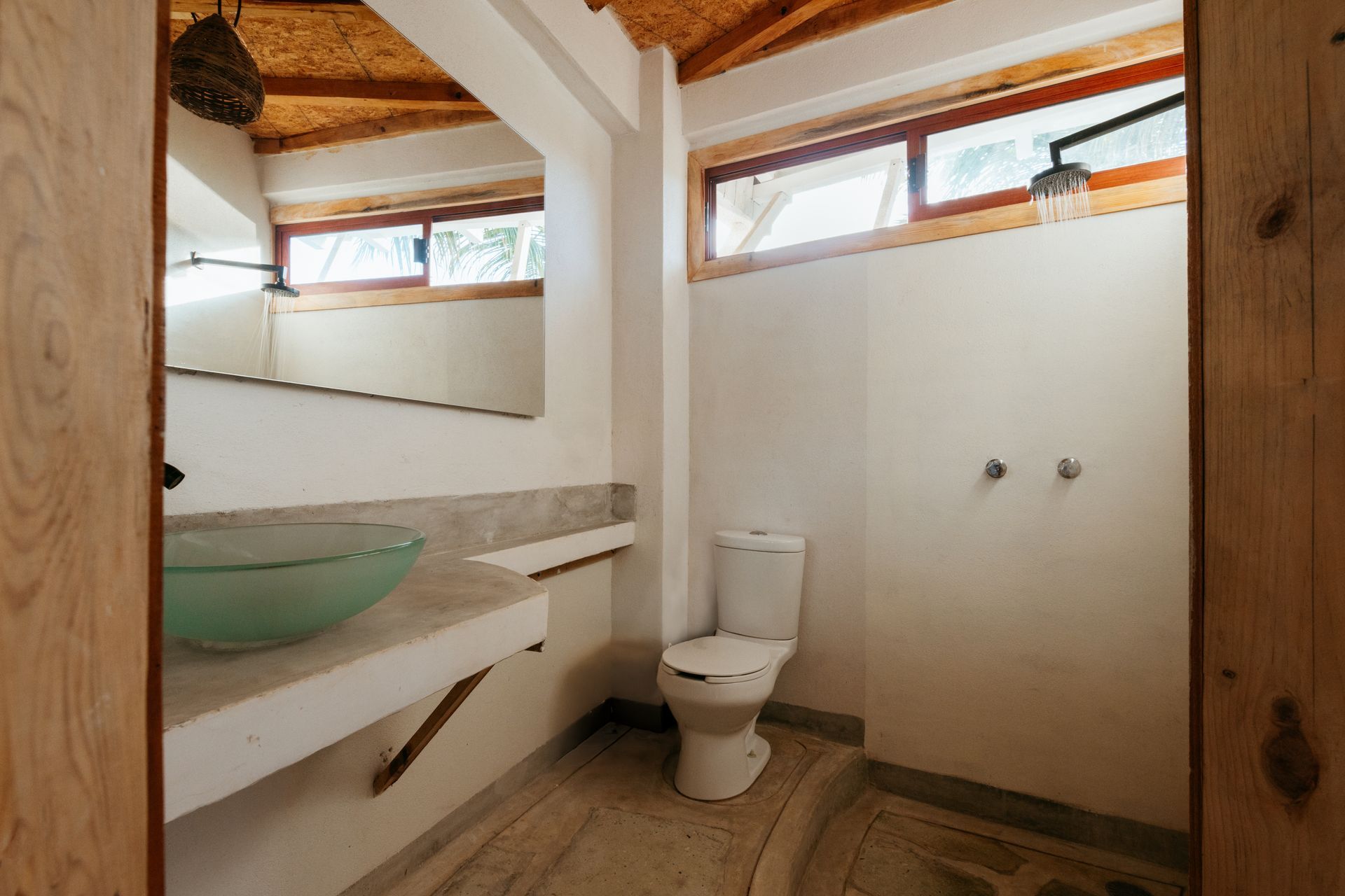 Baño con paredes blancas, inodoro, lavabo, espejo y ducha bajo vigas de madera en posada del arquitecto en mazunte, oaxaca