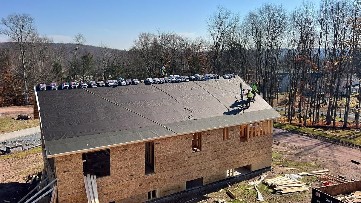 Construction of a house with a partially shingled roof, surrounded by trees, on a sunny day.