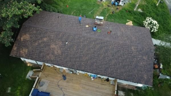 Aerial view of roof with workers. Brown shingles, green grass, and a wooden deck.