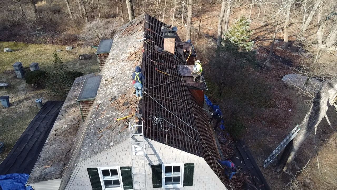 Roof being repaired; workers on a partially shingled roof with a ladder and view of the ground.