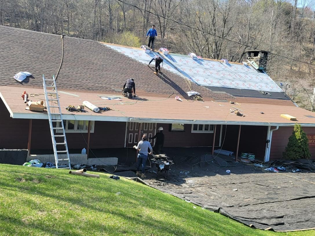 Roofers replacing a roof on a red house. Several workers are on the roof and on the ground. Green grass surrounds the house.