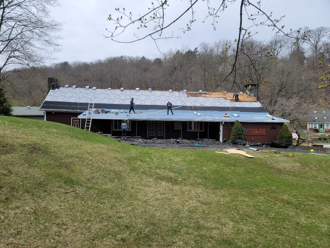 House with partially replaced roof, workers visible, green lawn in foreground, trees in background.