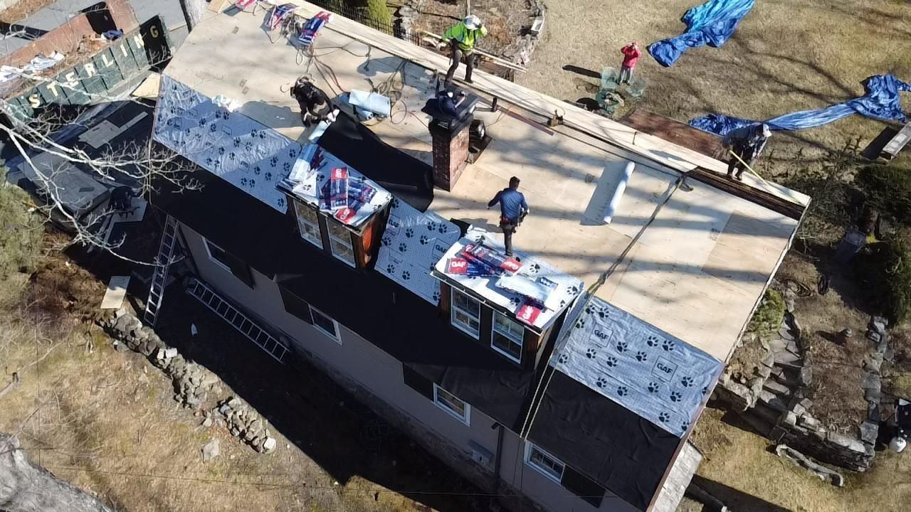 Roofing crew working on a two-story house with dark shingles, light wood, and blue tarps.