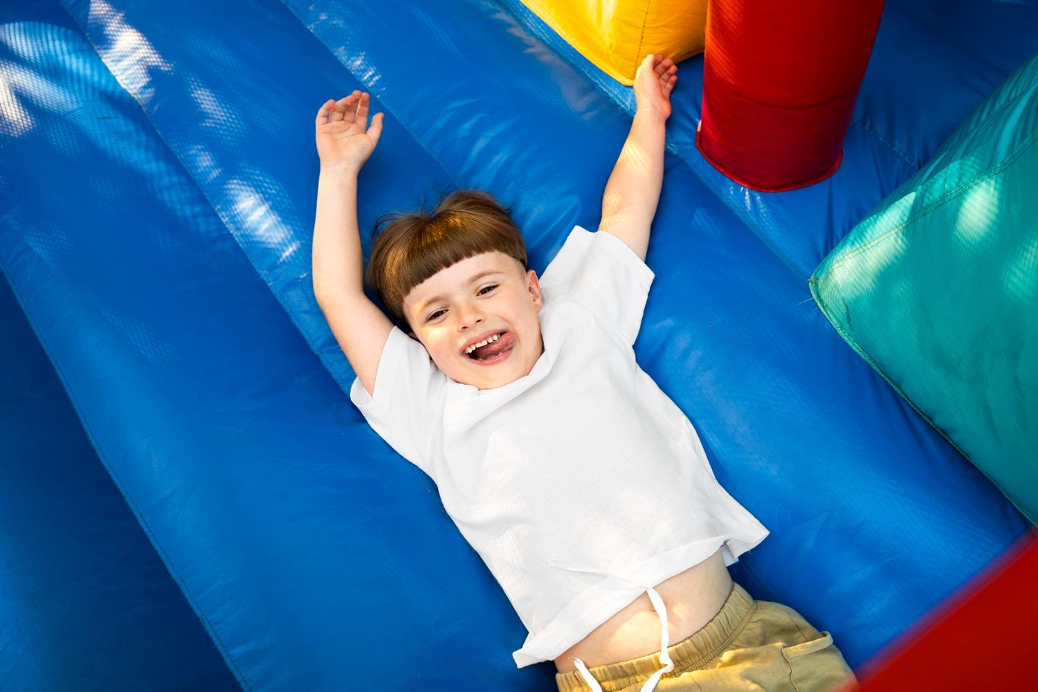 Child smiling on a blue inflatable slide with arms raised, wearing a white shirt and gold pants.