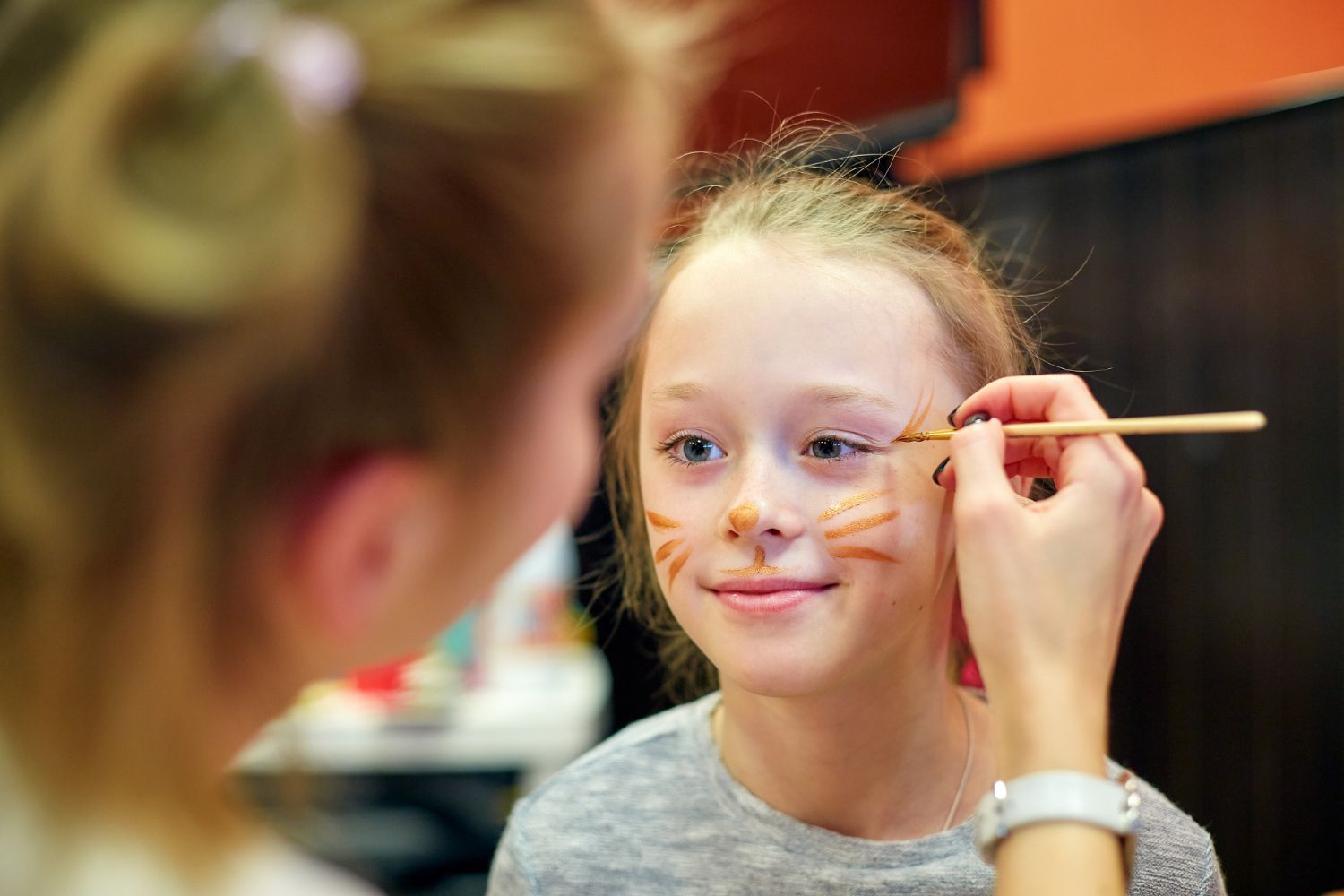 Child having face paint applied by an adult with a brush in a warm indoor setting