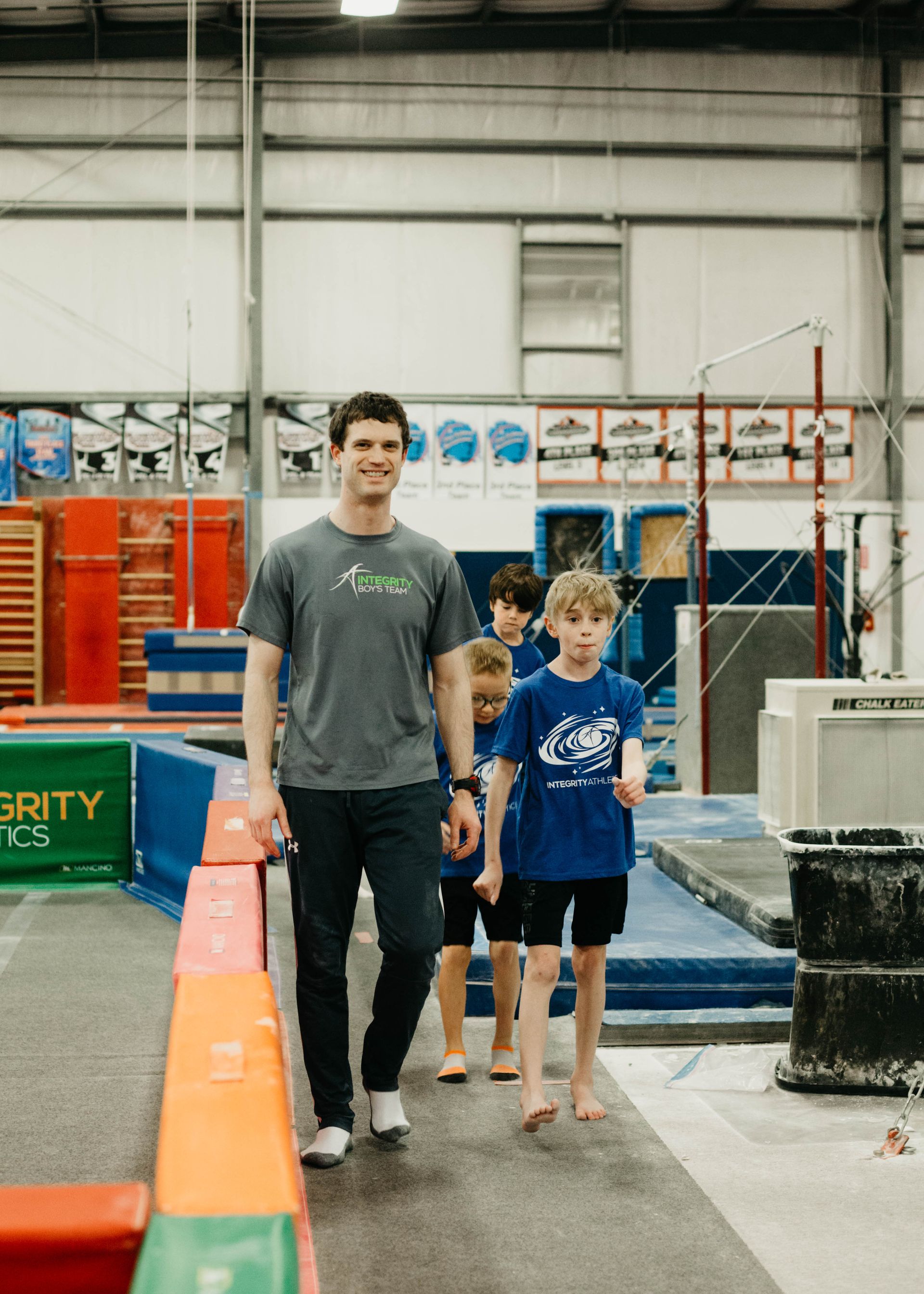 A man and three children are walking across a balance beam in a gym.