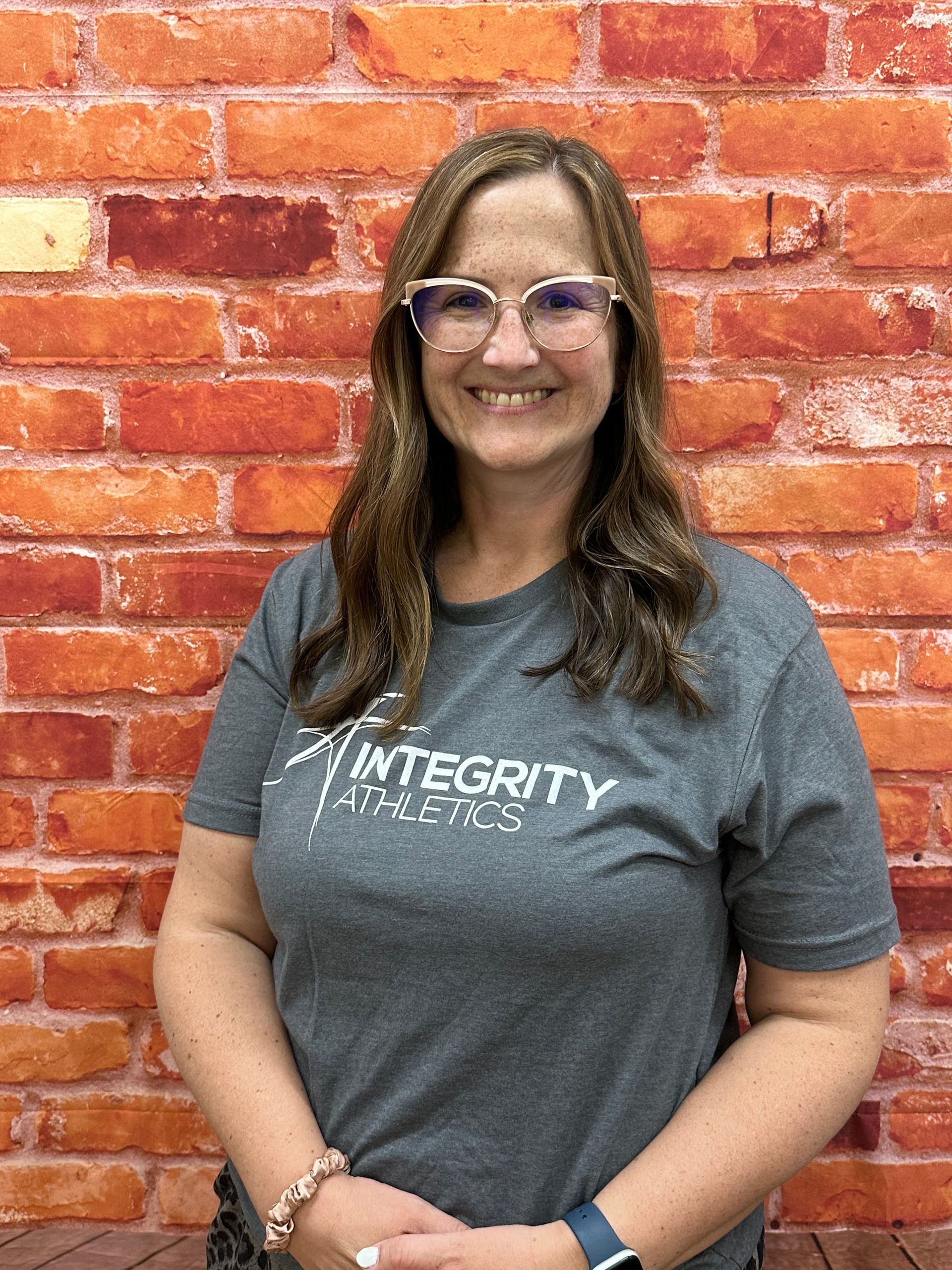 Smiling person in gray Integrity shirt standing in front of a red brick wall