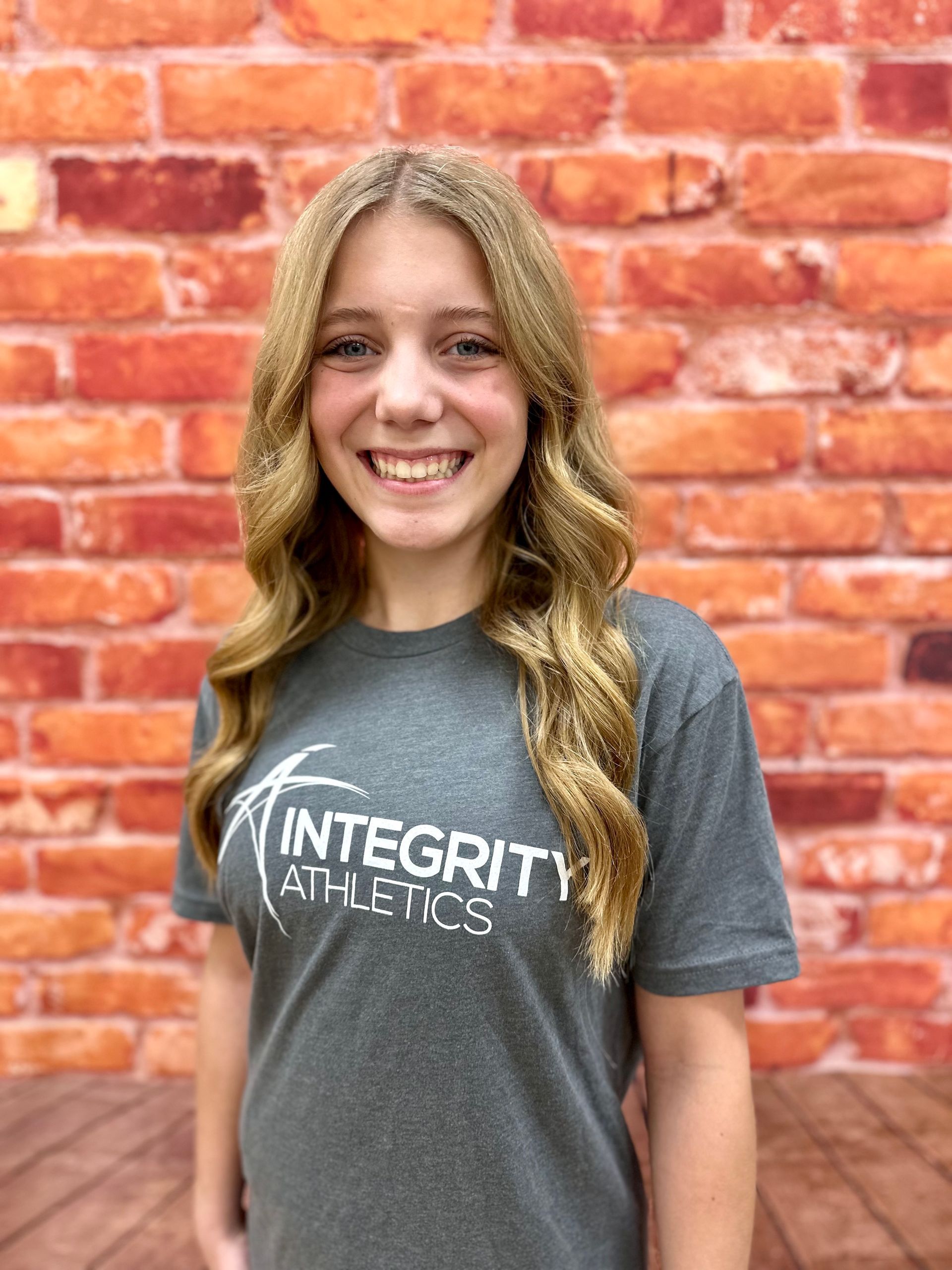Smiling person in a gray “Integrity Athletics” T-shirt standing in front of a red brick wall