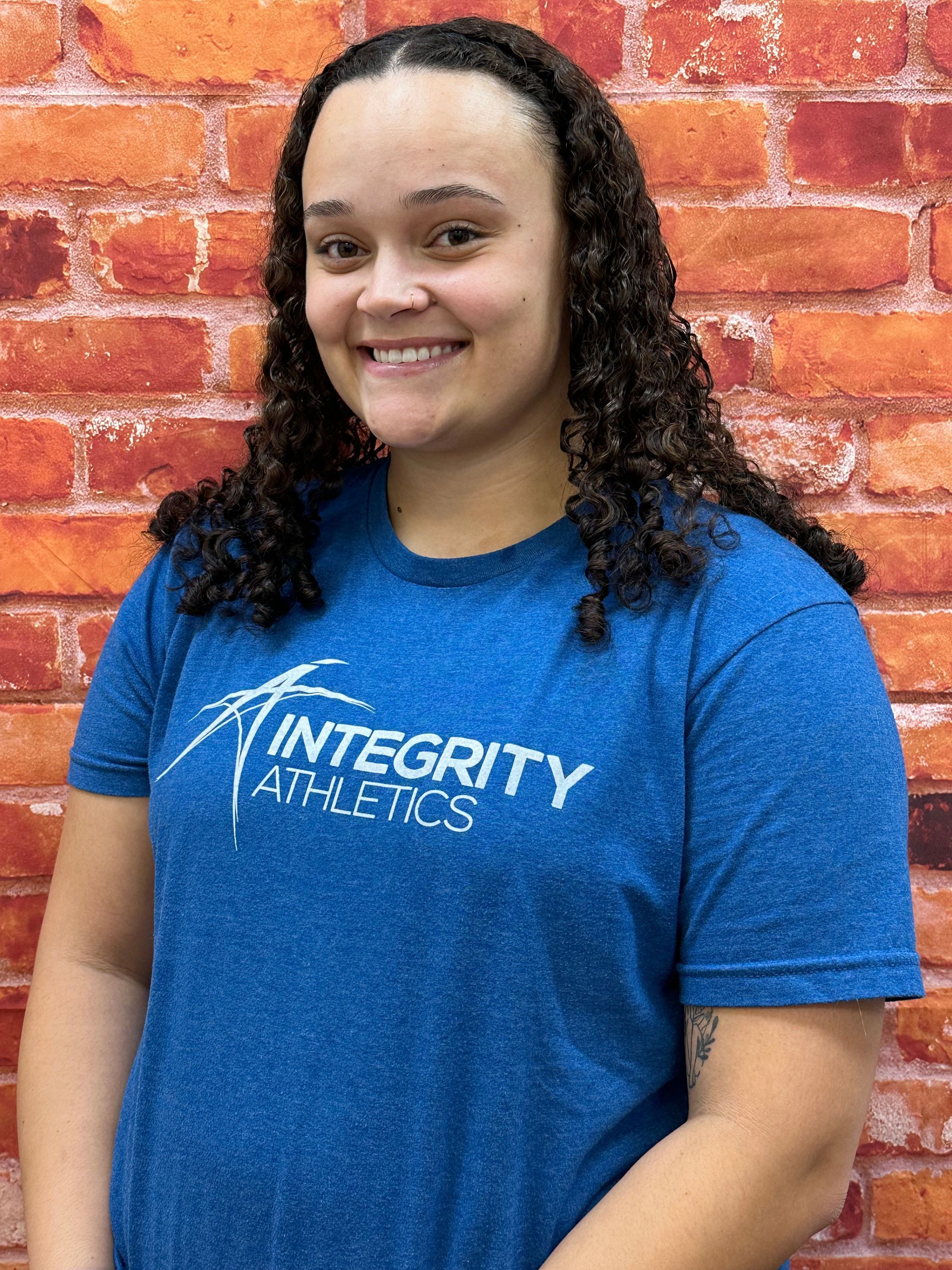 Smiling person in blue Integrity Athletics T-shirt standing in front of a brick wall.