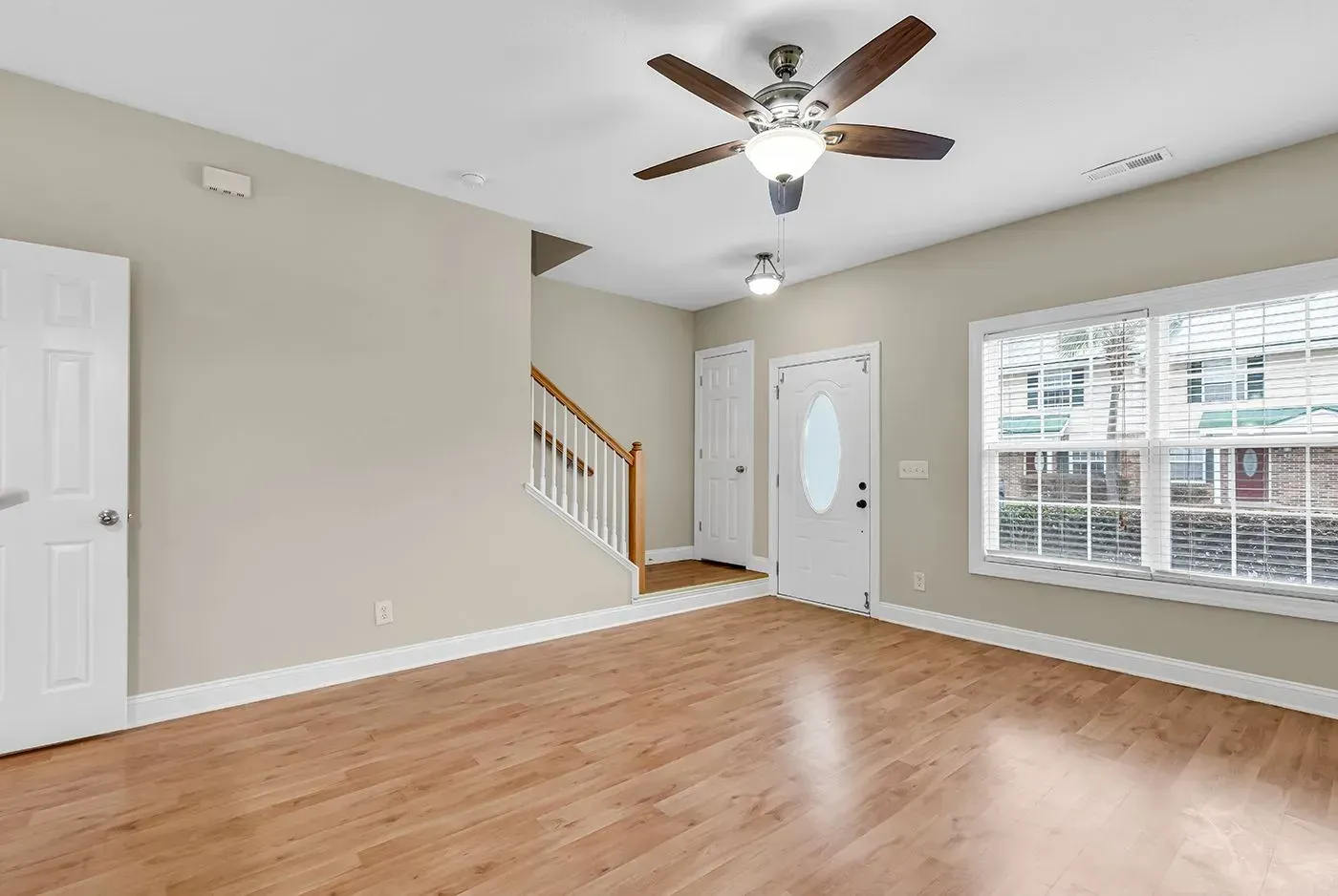 Empty living room with wood floors, beige walls, ceiling fan, staircase, and large window.
