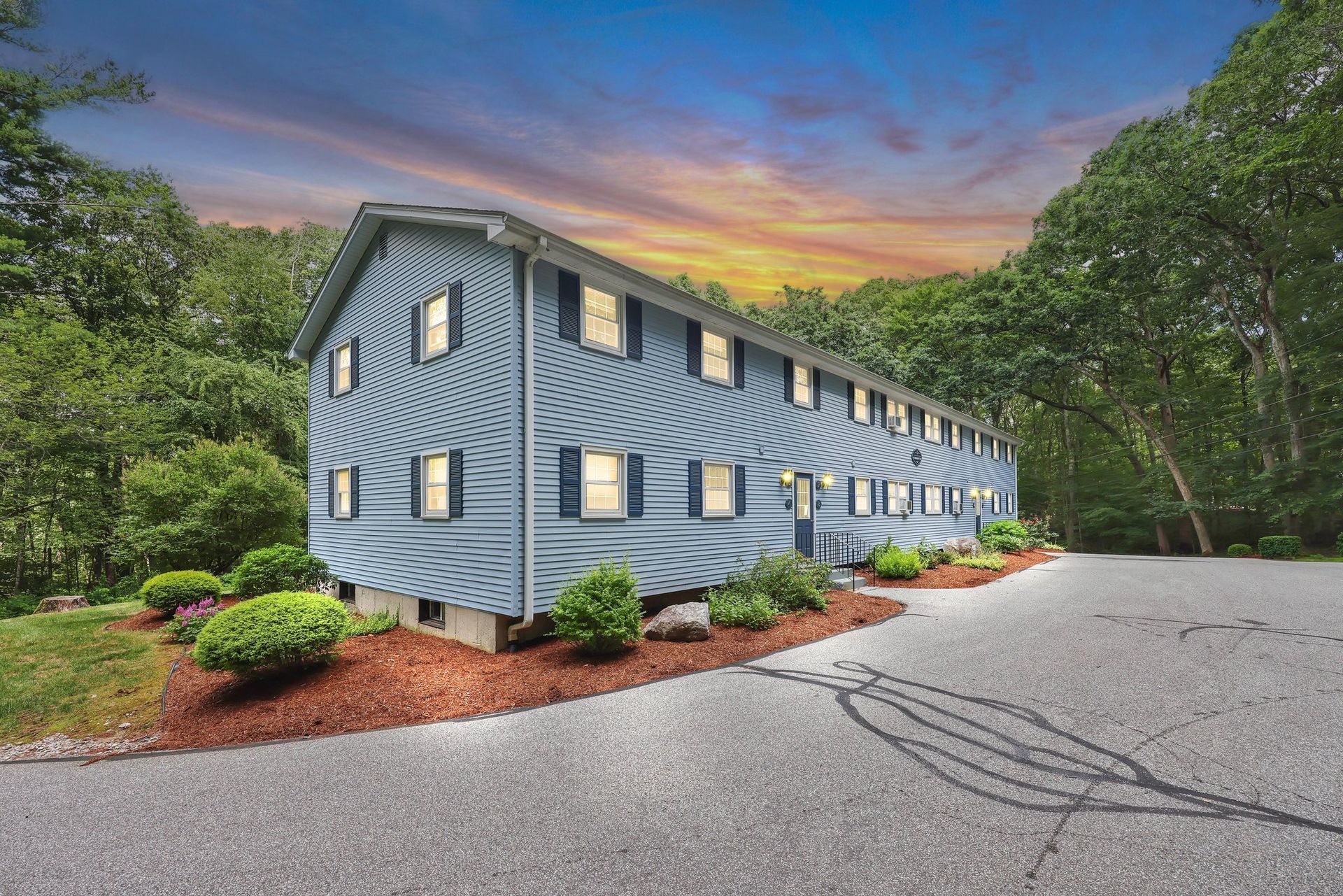 Blue multi-story building beside a curved driveway, surrounded by trees at sunset