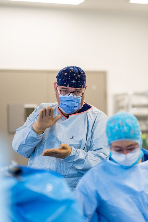 Surgeons in blue scrubs and masks in an operating room, one holding a hand up while another stands nearby