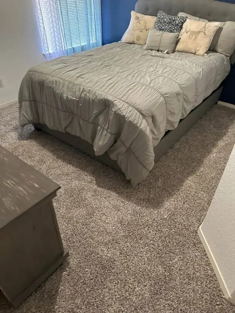 Small bedroom with a neatly made bed, gray comforter, pillows, and a carpeted floor near a window.