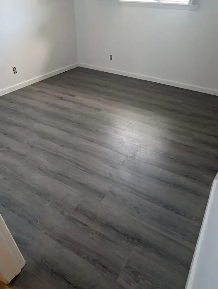 Empty room with gray wood-look flooring and white walls, lit by a small window.