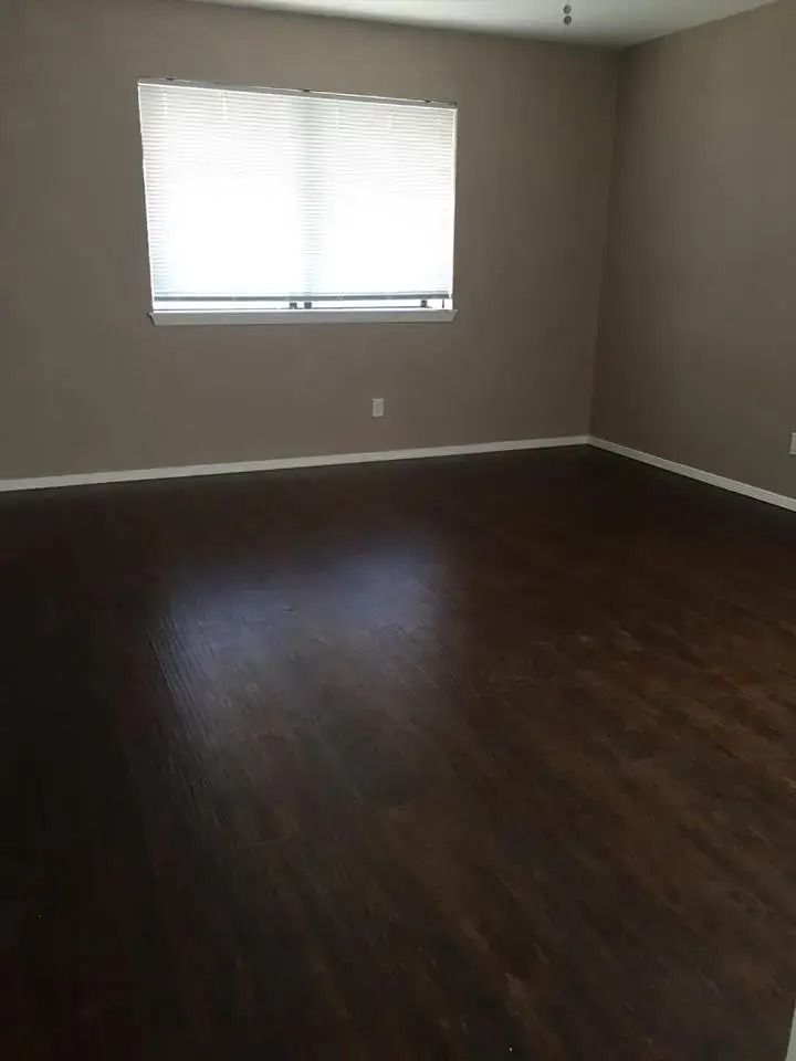Empty room with dark wood floor, beige walls, and a bright window with blinds.