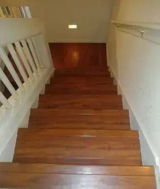Wooden staircase descending to a lower landing with white walls and railing, lit by a small wall light.