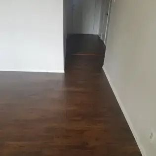 Empty room with dark wood floor, white walls, and a hallway in the back