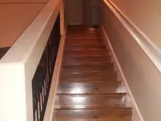 Narrow wooden staircase with white walls and black railing leading upward to a closed door.