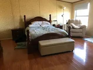 Bedroom with a wooden bed, beige ottoman, and armchair on a hardwood floor