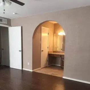 Empty room with dark wood floors and an arched doorway leading to a warmly lit bathroom.
