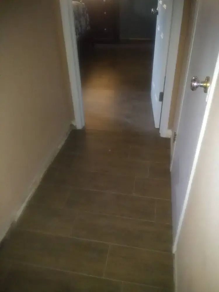 Narrow hallway with brown tile floor, beige walls, and white door frames leading into a darker room