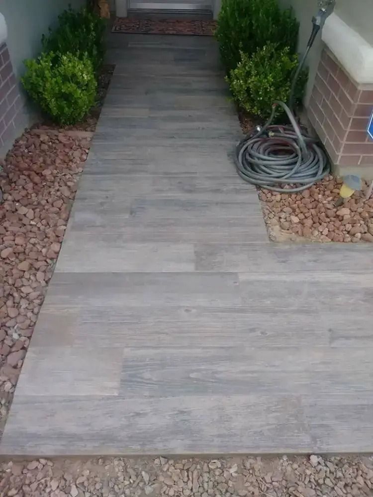 Front walkway with large gray tiles, potted shrubs, and a coiled garden hose beside brick and pebble borders