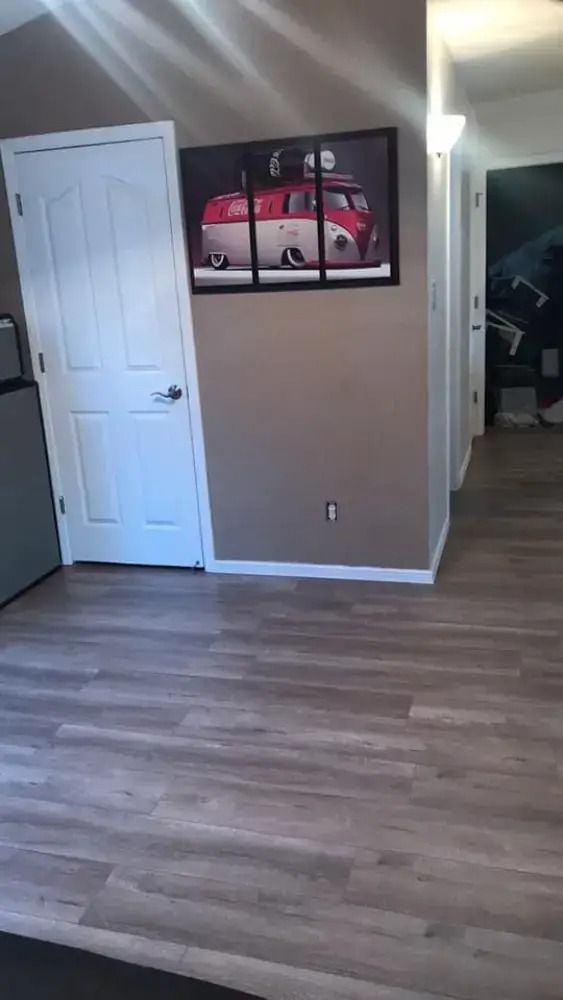 Empty room with light wood flooring, white door, and framed car artwork on a taupe wall.