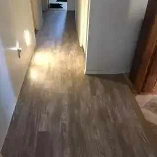 Narrow hallway with wood-look flooring and white walls, lit by sunlight from a room at the end.
