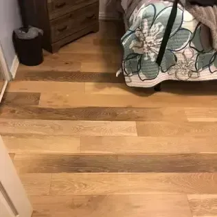 Wooden floor in a bedroom, with a dresser, trash can, and a bed with patterned bedding.