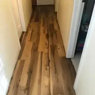 Narrow hallway with light walls, white door frames, and a wood plank floor with dark grain patterns.