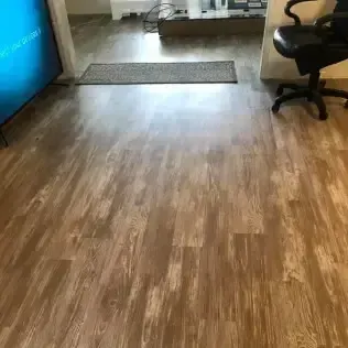 Empty living room with wood flooring, a rug by the doorway, and a black office chair on the right.