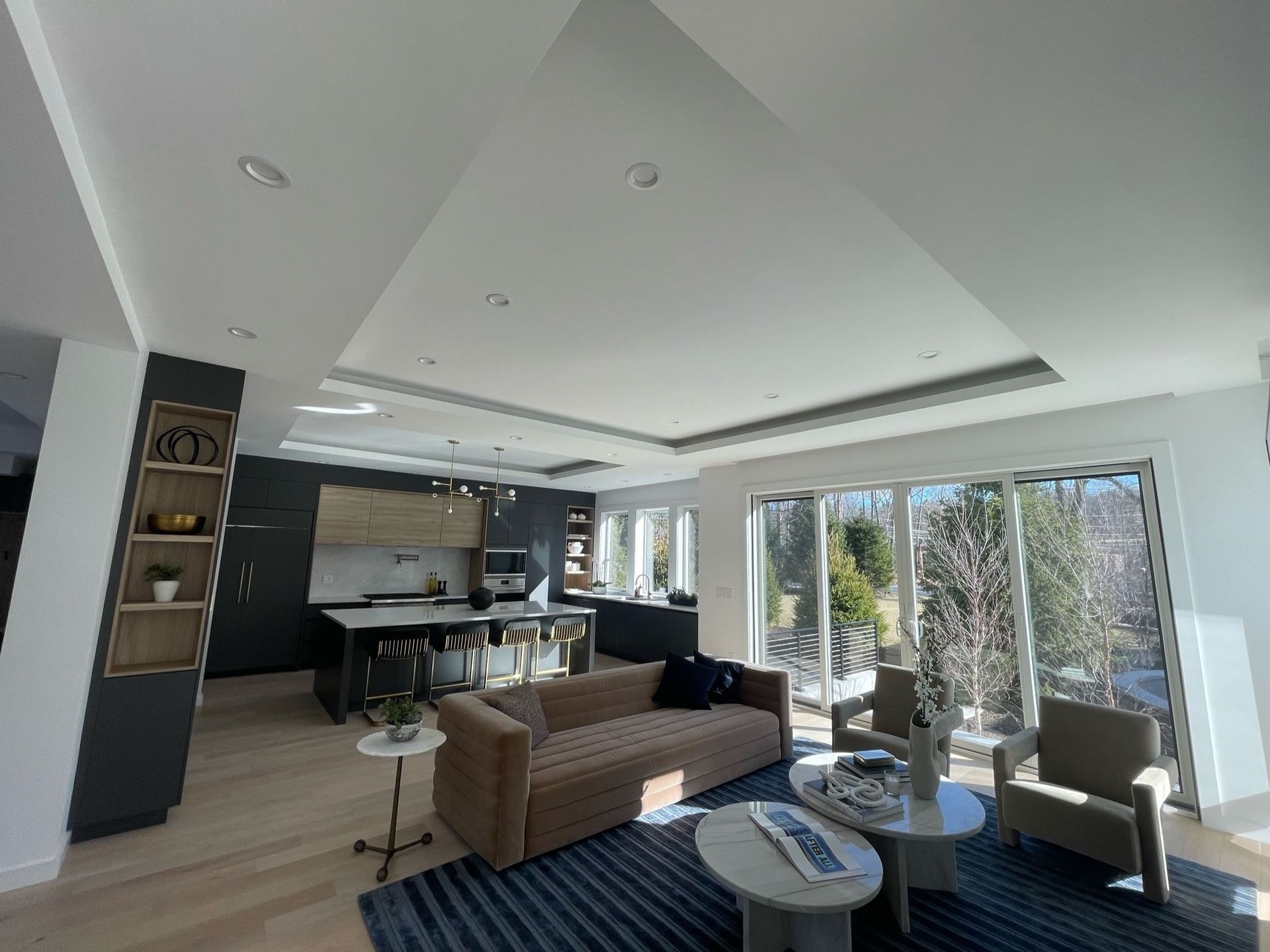 Bright modern open-plan living room with sofa, coffee table, kitchen, and large windows overlooking greenery