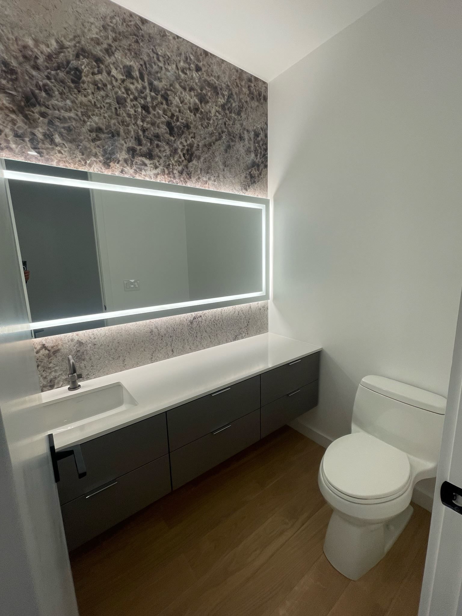 Modern bathroom with illuminated mirror, dark vanity, and toilet near a white wall