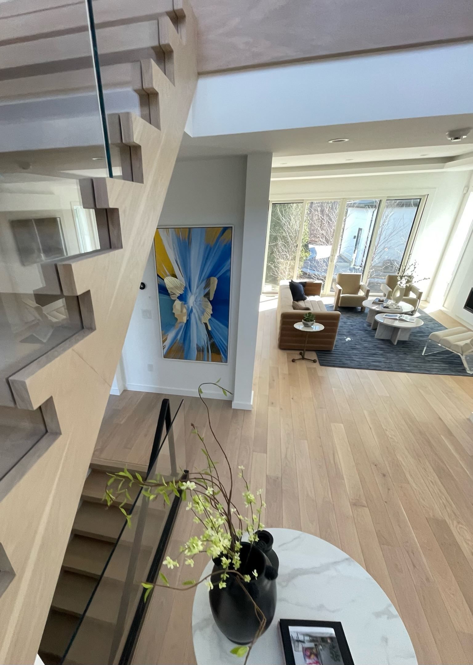 Modern living room with staircase, abstract wall art, and a dining area by large windows.