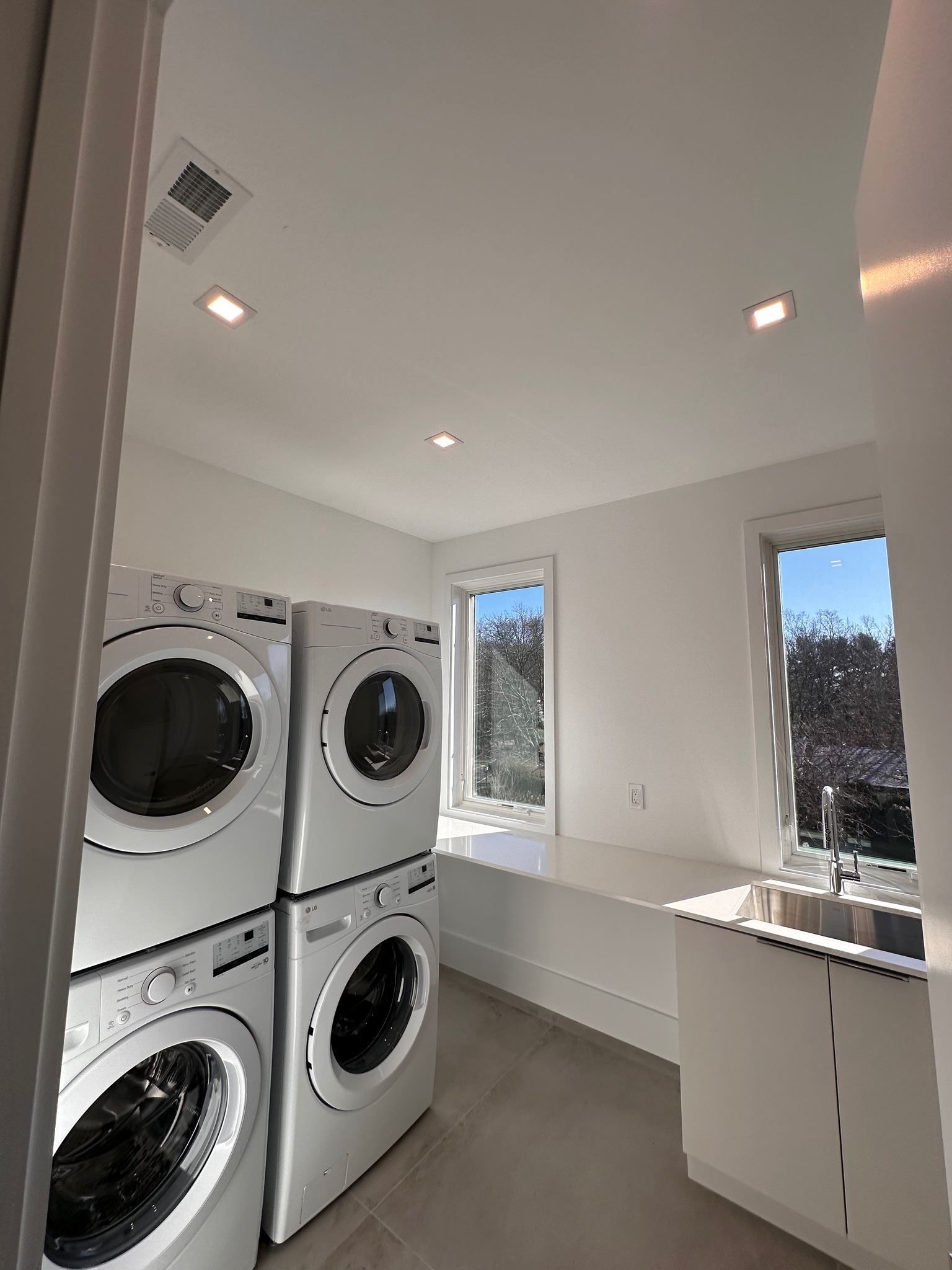 Laundry room with stacked washer and dryer units, white cabinets, and windows overlooking trees.