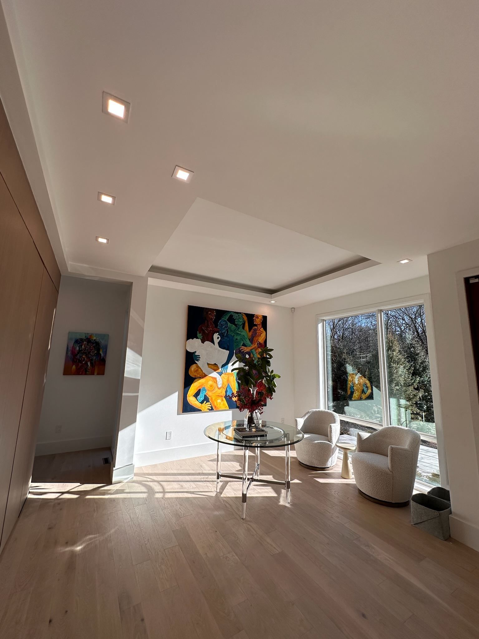 Modern living room with skylight, white chairs, colorful wall art, and large windows overlooking trees