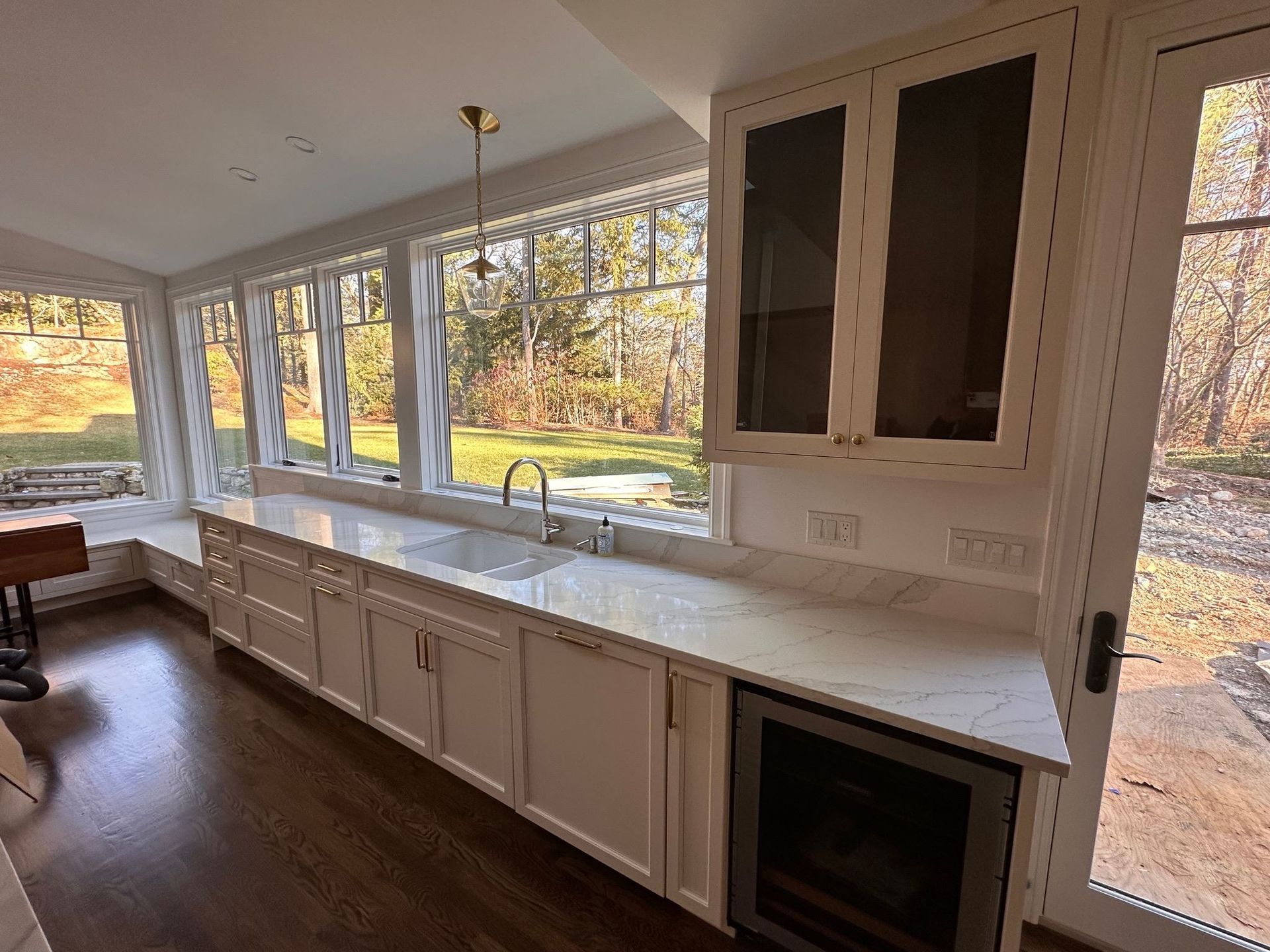 Bright kitchen with long white counters, many windows, and dark wood floors overlooking trees outside