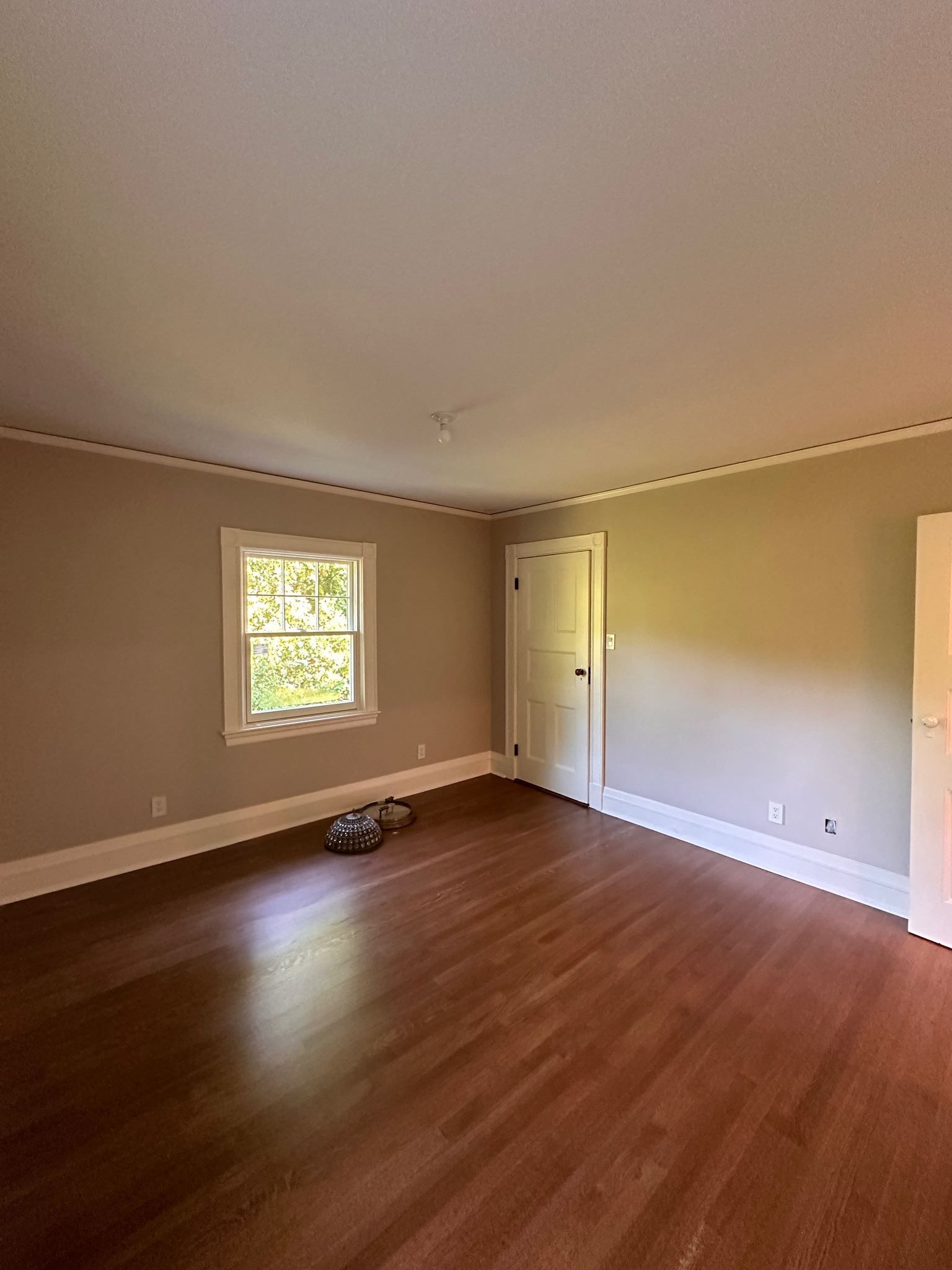 Empty room with hardwood floors, beige walls, a window, and a white door.